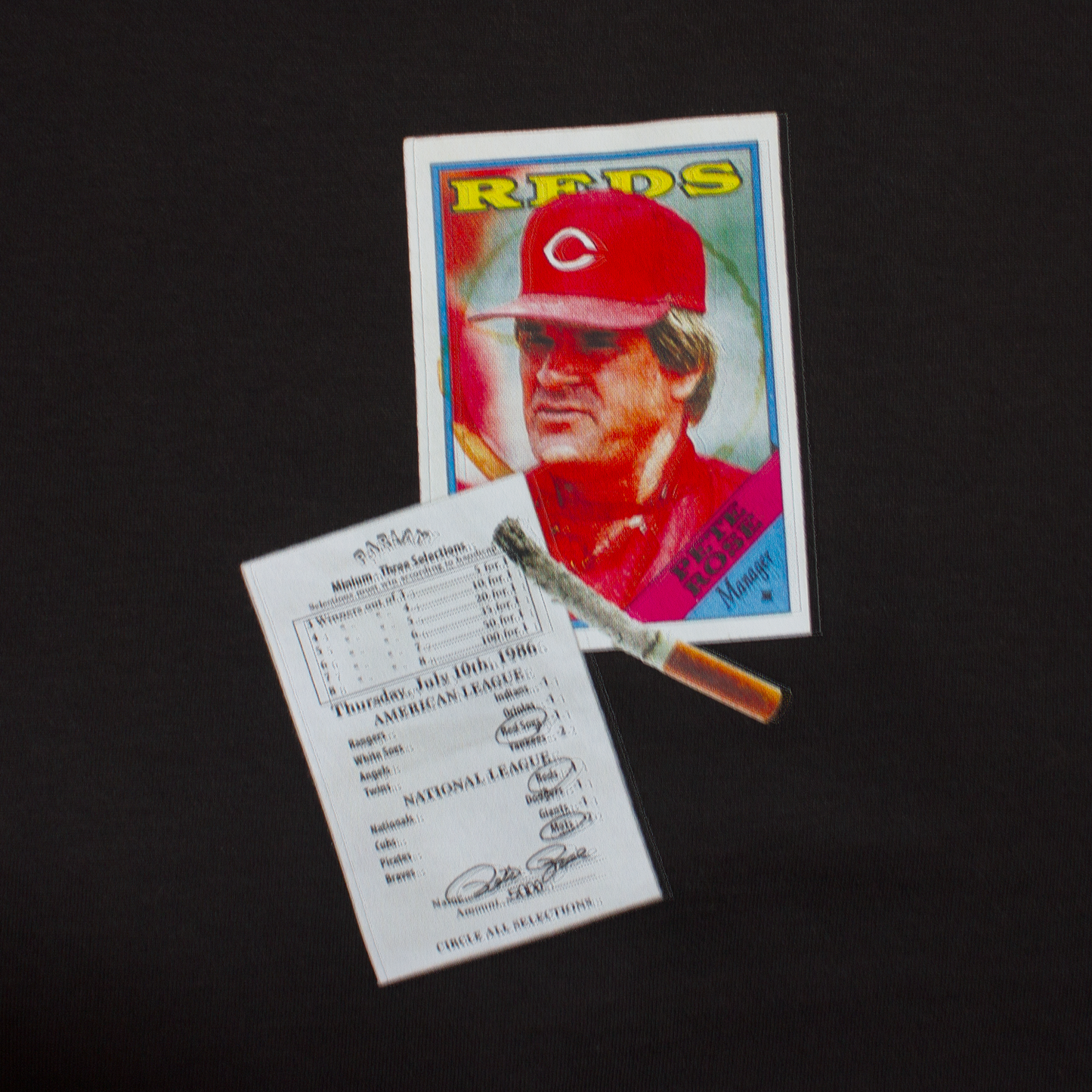 close up of a t-shirt with a baseball card of Pete Rose along with a burning cigarette and a betting slip filled out by Pete Rose betting on the Cincinnati Reds 